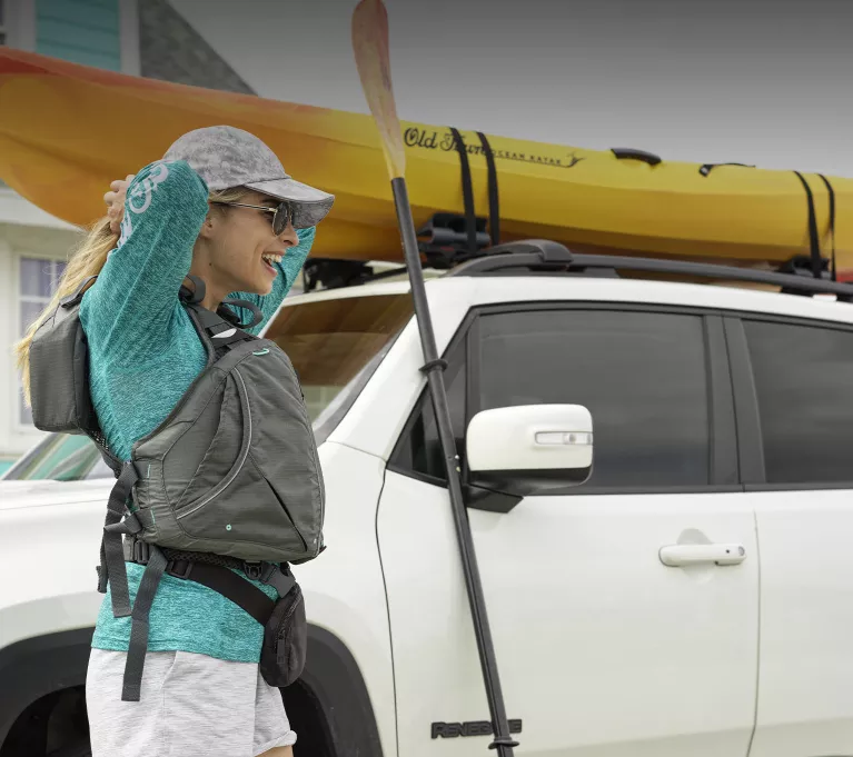 A woman adjusts her life jacket while standing next to her car and kayak. 