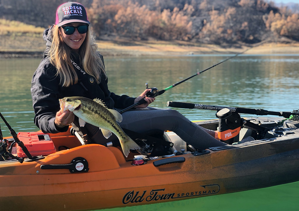 Annie Nagel holds up a medium size largemouth bass caught on a fly rod in an orange fishing kayak. 