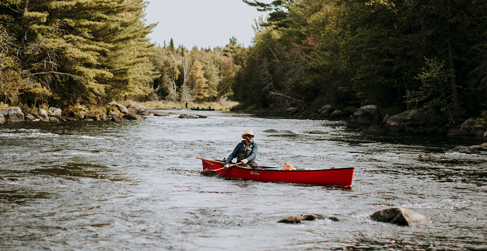 A man in a cowboy hat paddles a red Old Town canoe down a small rapid on the St. Croix river in Maine. 