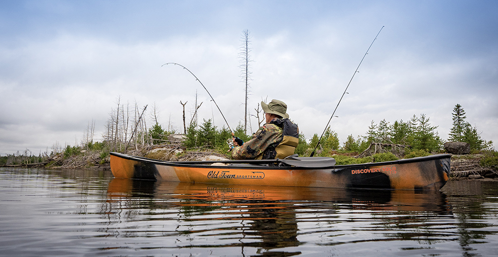 A man casts a fishing rod from an orange and black Old Town Sportsman Discovery Solo 119 canoe. 