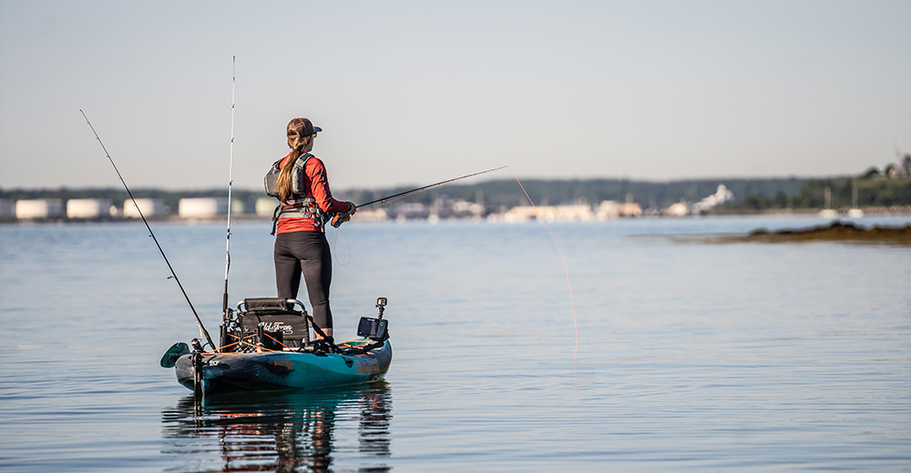 Christi Holmes stands and casts a saltwater plug from a teal fishing kayak on a calm saltwater bay. 