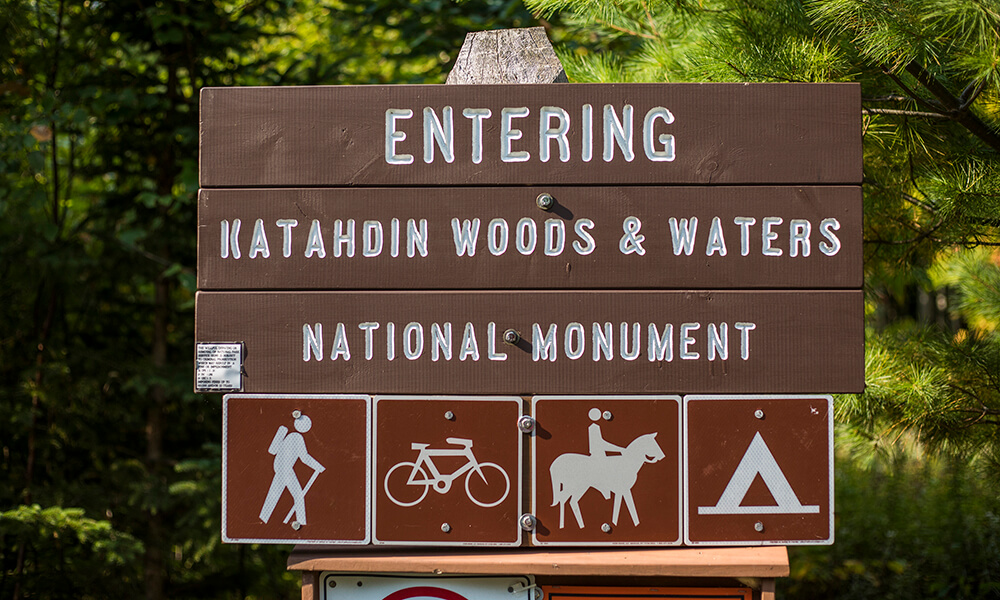 Photograph focused on the brown park entrance sign, reading Entering Katahdin Woods &amp; Waters National Monument, with icons depicting hiking, cycling, horse riding, and camping. 