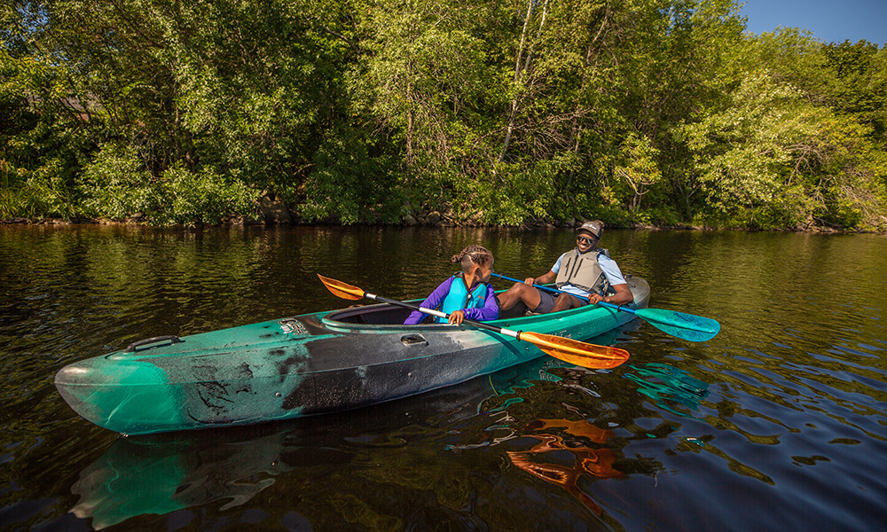 Photograph showing an african american man and his young daughter paddling a teal tandem kayak on a calm river on a sunny day. 