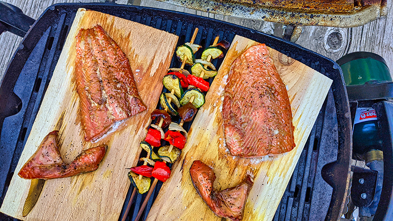 Two freshly cooked salmon filets rest on cedar planks atop a grill, with two kebabs loaded with grilled vegetables between the two planks. 