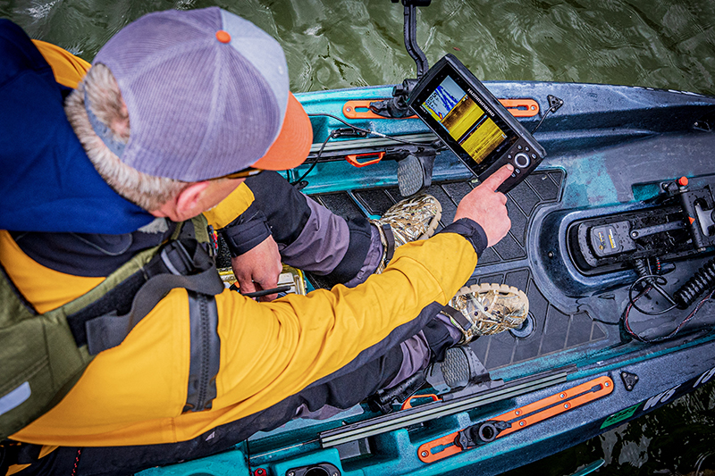 A kayak angler leans over the cockpit of their kayak to press a button on their fish finder.