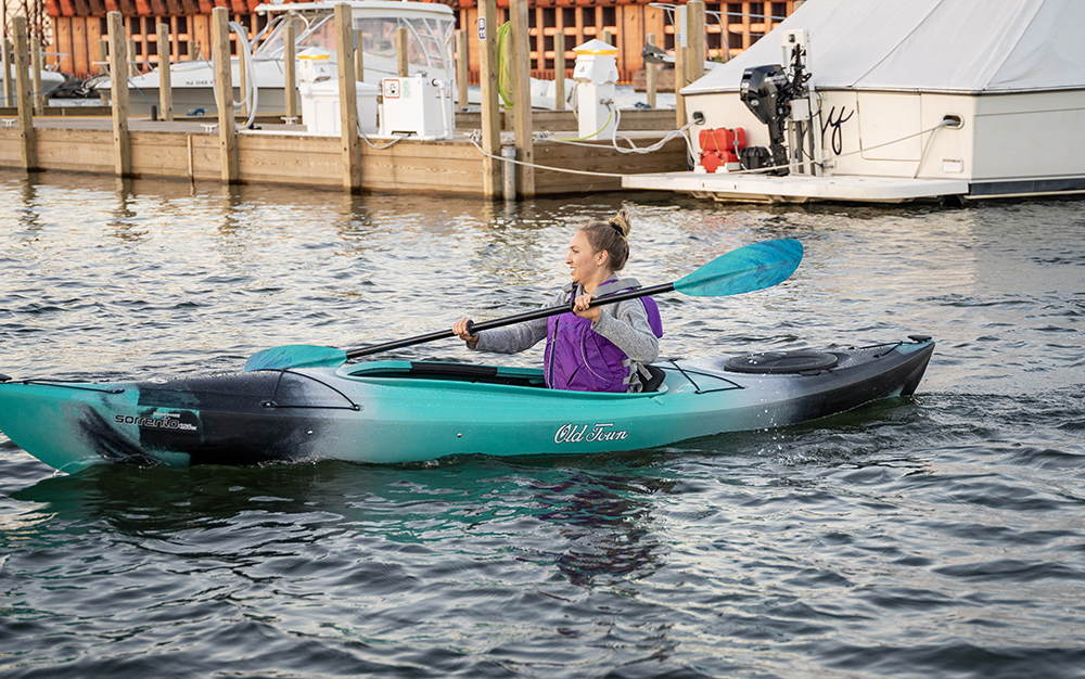 A woman paddles a teal and black Old Town Sorrento kayak on a calm harbor with boats in the background.