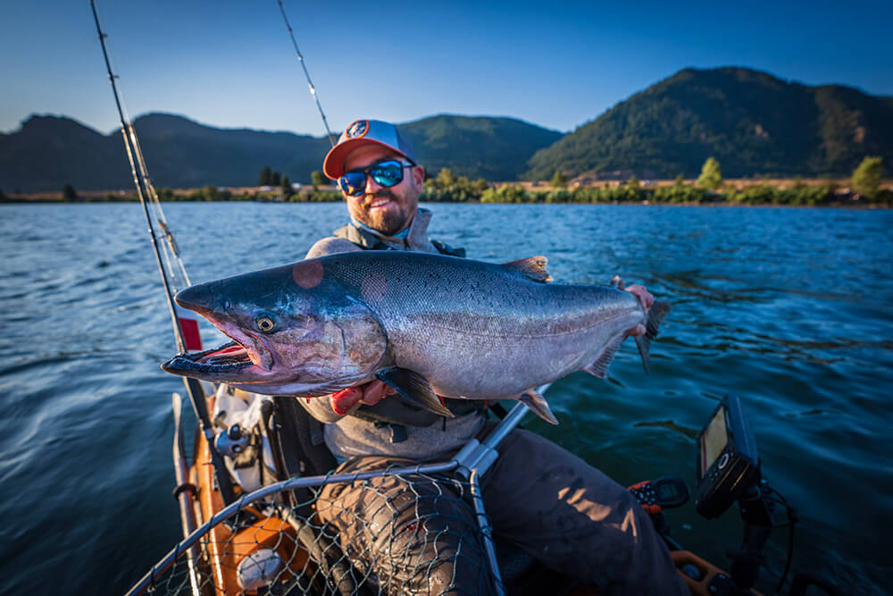 A bearded man lifts a large salmon up while seated in a fishing kayak. 