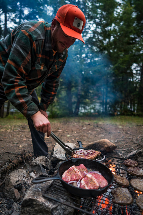 A man cooks steaks in a cast iron skillet on a campfire. 