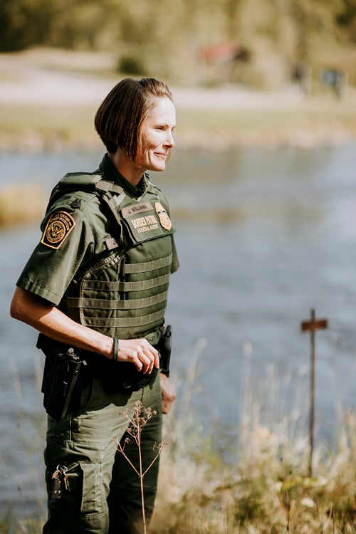 A border patrol agent stands near the river and smiles.