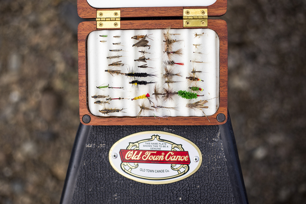 A close-up photograph of a fly box sitting on the bow of an Old Town canoe with traditional hand-tied flies inside the box. 