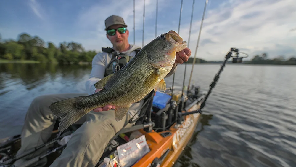 Casey Reed lifts a big largemouth bass with both hands while sitting in his AutoPilot 136 fishing kayak. 