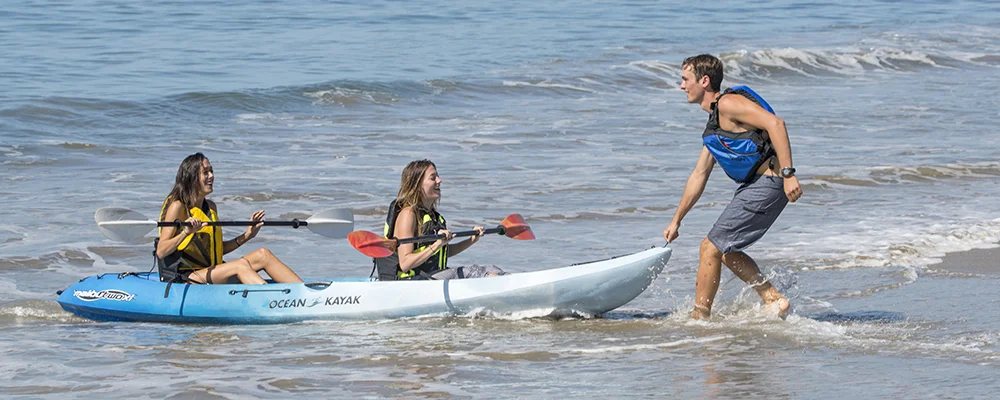 A man pulls two tandem kayakers onto the beach on a sunny day. 