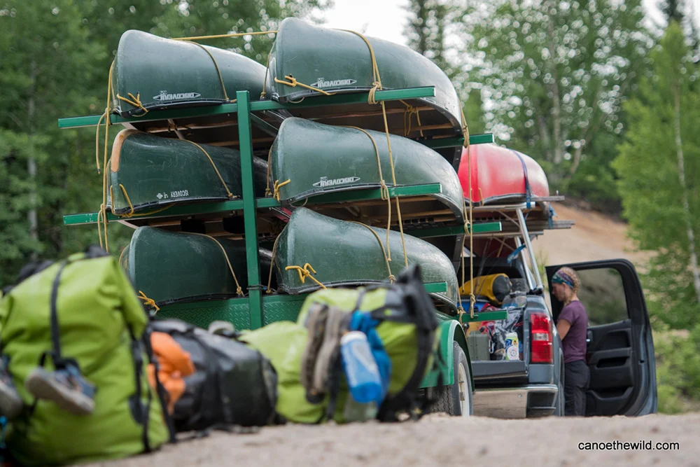 A stack of 9 canoes on a trailer attached to a truck, while a guide retrieves gear from the cab of the truck. 