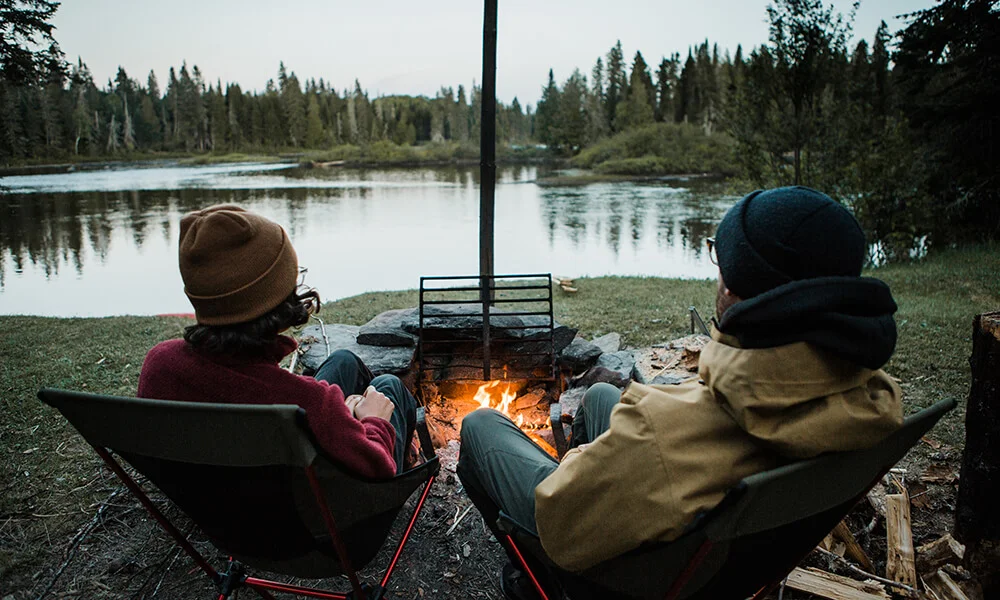 Photograph showing two campers from behind, seated at a remote wilderness campsite looking at a fire in a fire pit overlooking a calm river. 