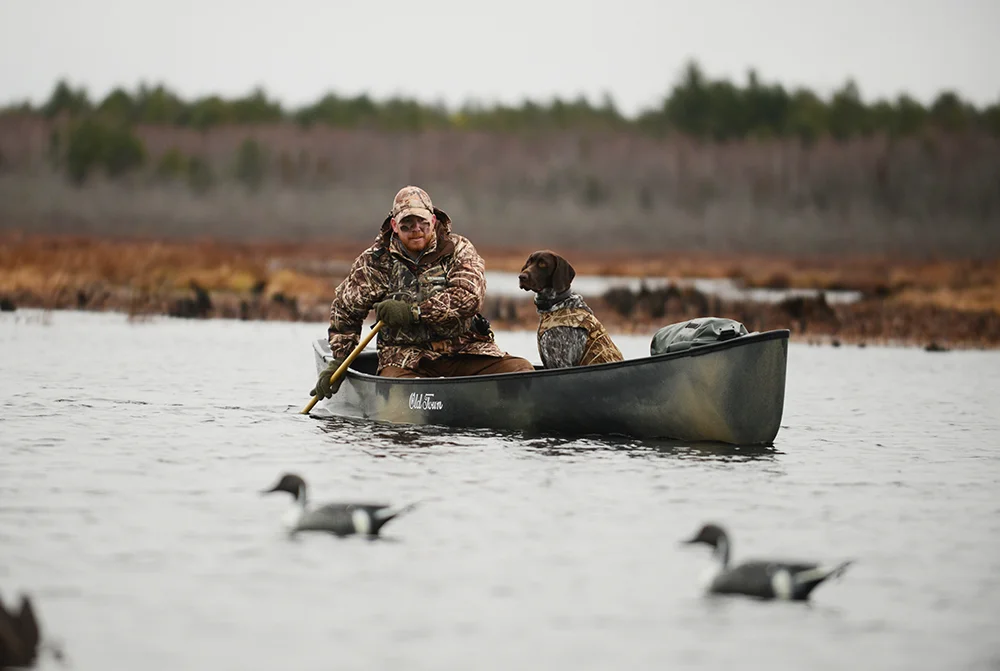 A hunter dressed in all camo paddles a canoe quietly on a cold winter morning, his dog sits in front of him wearing a camo life jacket.