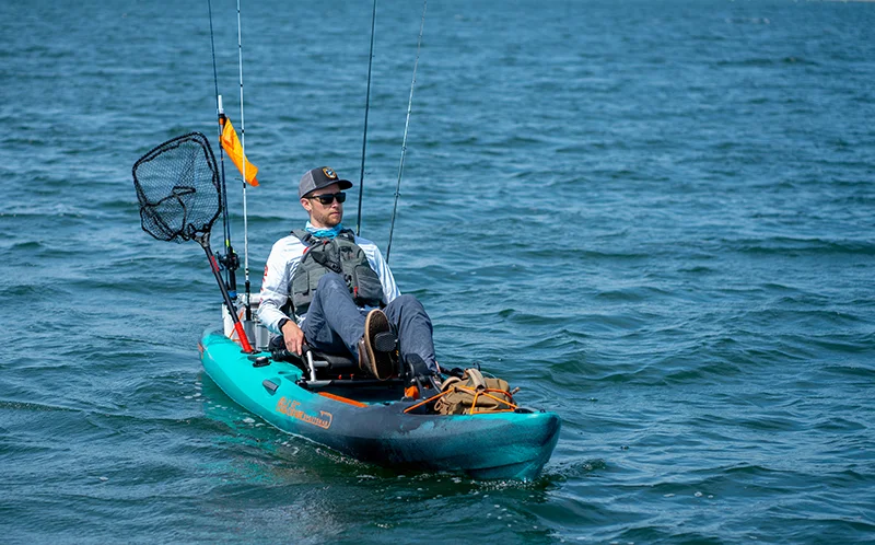 A male kayak angler pedals an Old Town Sportsman Salty PDL 120 on a clear sunny day, with an orange safety flag waving in the breeze. 