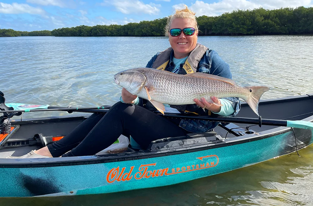 A blonde haired female kayak angler holds a large redfish while sitting in a teal solo canoe on a sunny calm day.