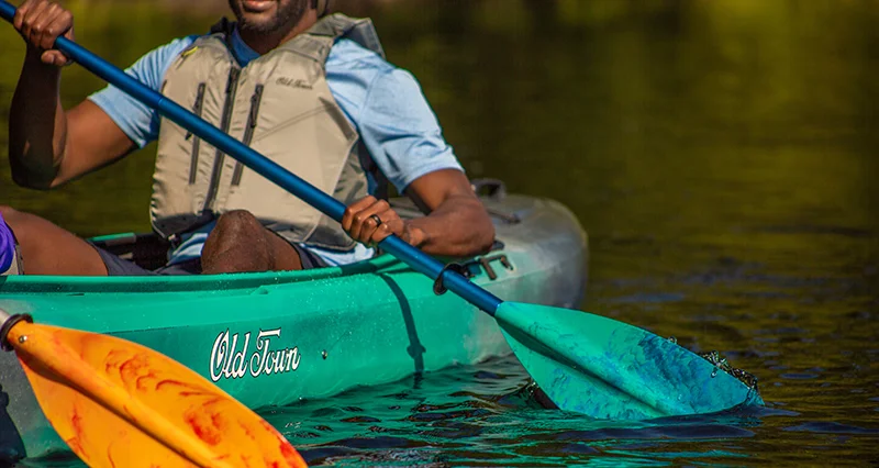 Close up image of a man wearing a gray life jacket paddling a tandem kayak with a brightly colored paddle. 