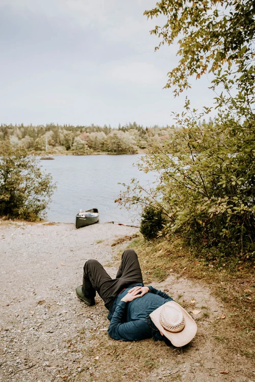 A man lays on the ground next to the river with his hat over his head. 