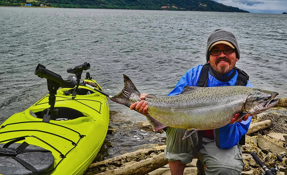 Tyler holds up a large Chilean King Salmon, kneeling next to his yellow kayak beached on a rocky shoreline. 