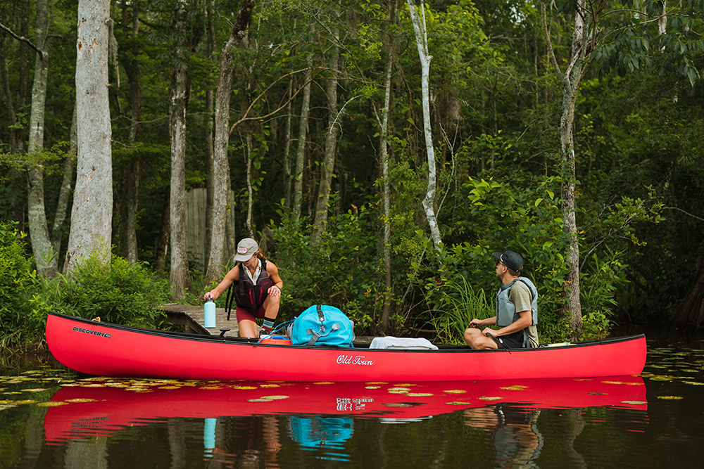 Two paddlers load their red Old Town canoe on the edge of the river, with the small elevated boardwalk to the tenting platform visible snaking through the dense woods in the background.