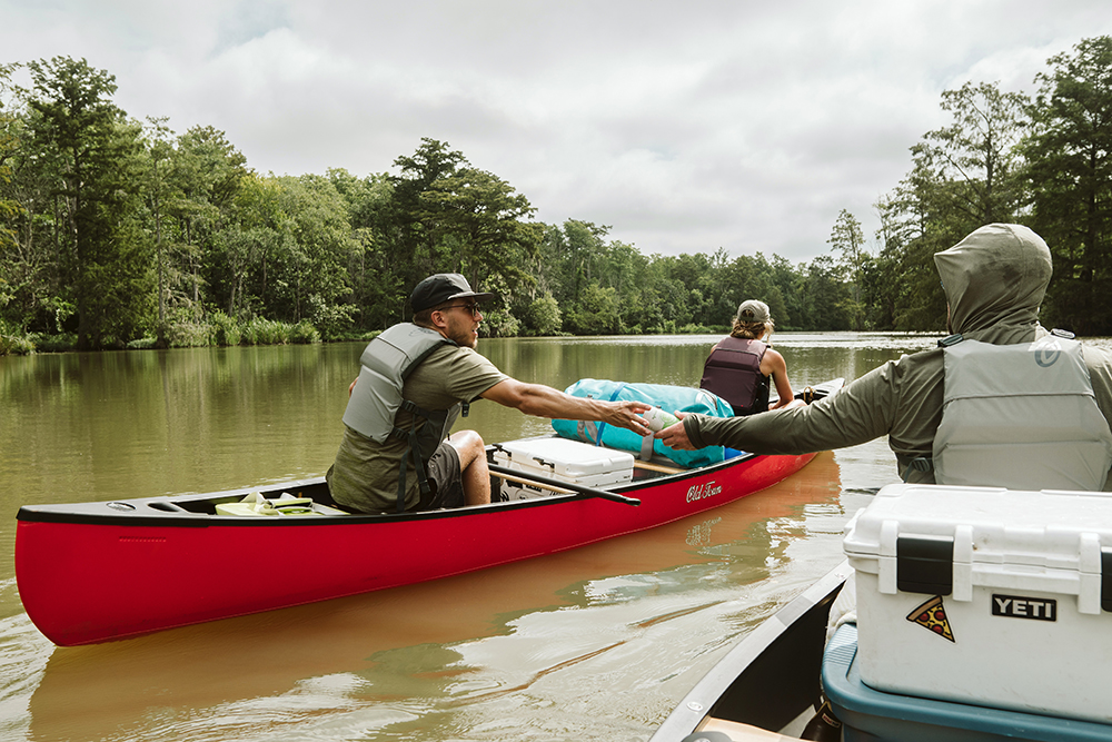 A hooded man passes a seltzer to a buddy in another canoe while paddling next to each other on the river. 