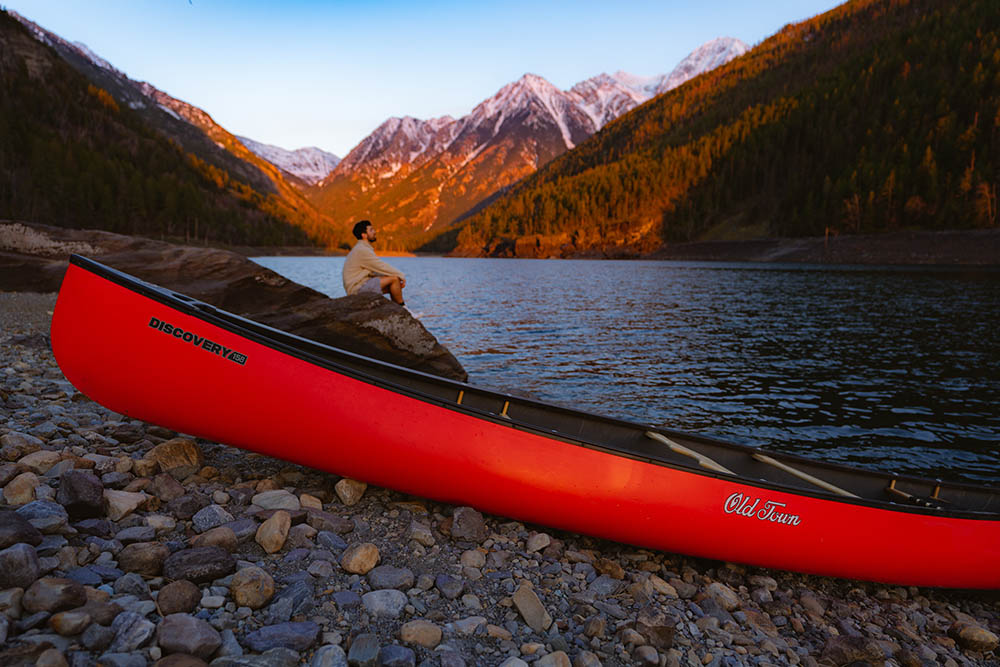 Man sitting by open water surrounded by mountains and Old Town Discovery 158 canoe resting on shore