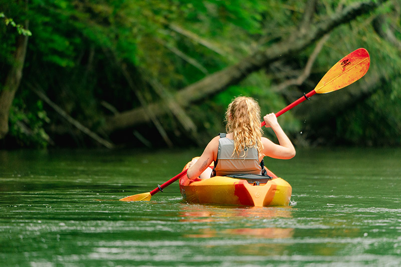 A picture of a female kayaker from behind paddling an orange kayak on a calm river with green river.