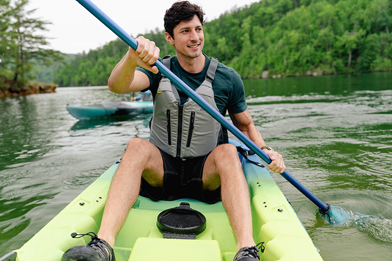A young man paddles a sit on top kayak while smiling, down a calm green river.