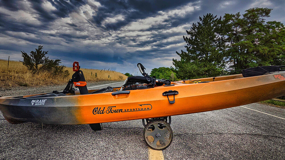 An orange and black fishing kayak sits on a bunk style Malone kayak cart with a dramatic cloudy sky in the background.