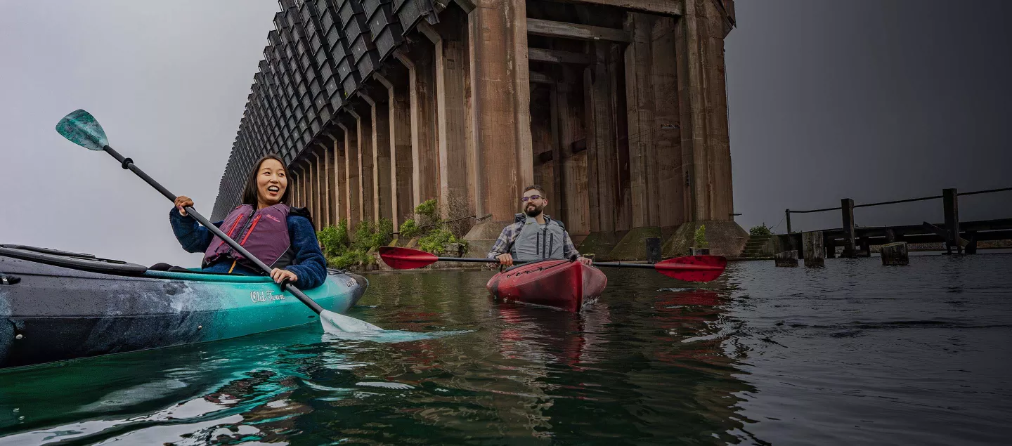 Two kayakers paddle on a calm bay on an overcast day.