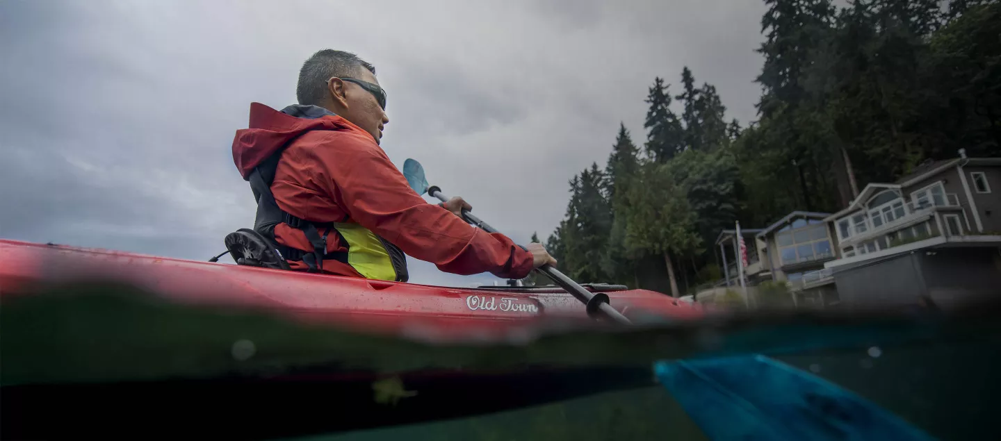 A man paddles a red Old Town kayak in deep green water.