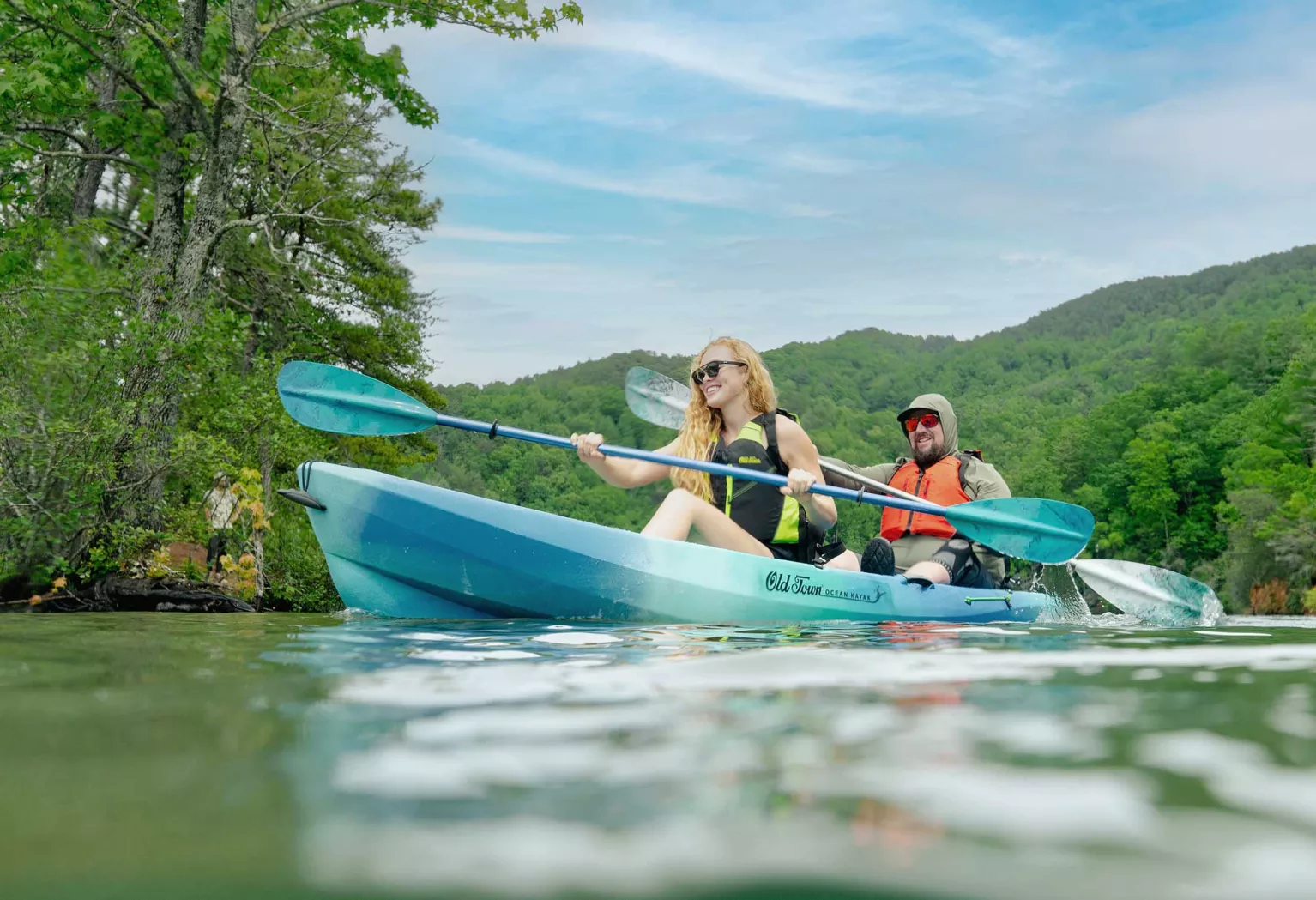 A man and woman paddle a teal tandem kayak on a murky but calm lake.