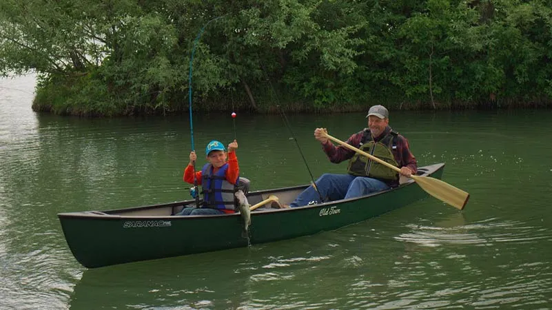 Man with paddle in hand and child holding up a caught fish in the Old Town Saranac canoe