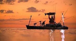 A kayak angler holds up a large king salmon, only a silhouette is visible against the dark orange sunrise.